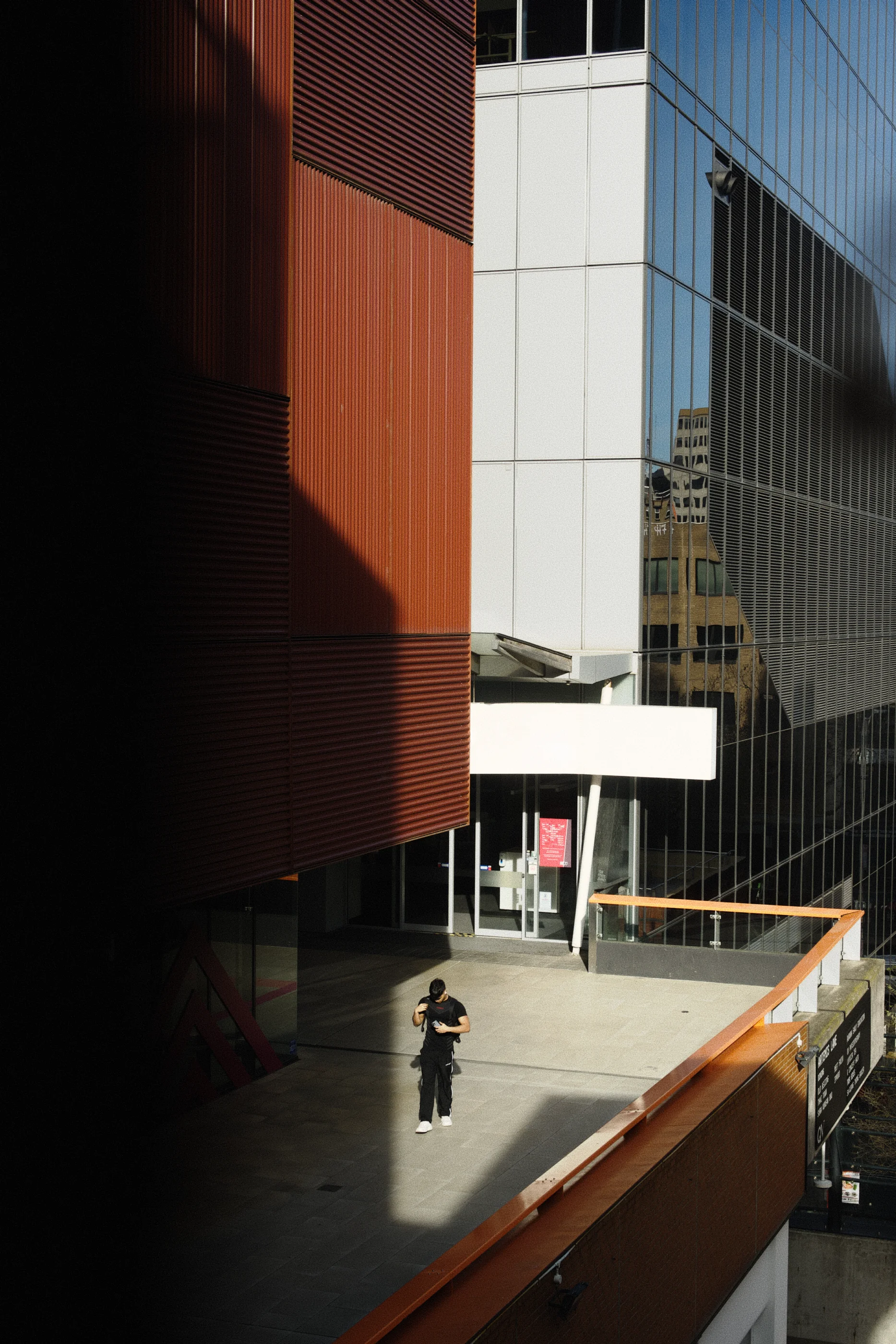 City buildings in a modern architecture. One in the foreground is cladded in red colorbond, one in the background has a glass facade appearing blue. In front of the red building is a terrace that is partially lit by sunlight. I male person dressed in black, is walking in the sunlit part.