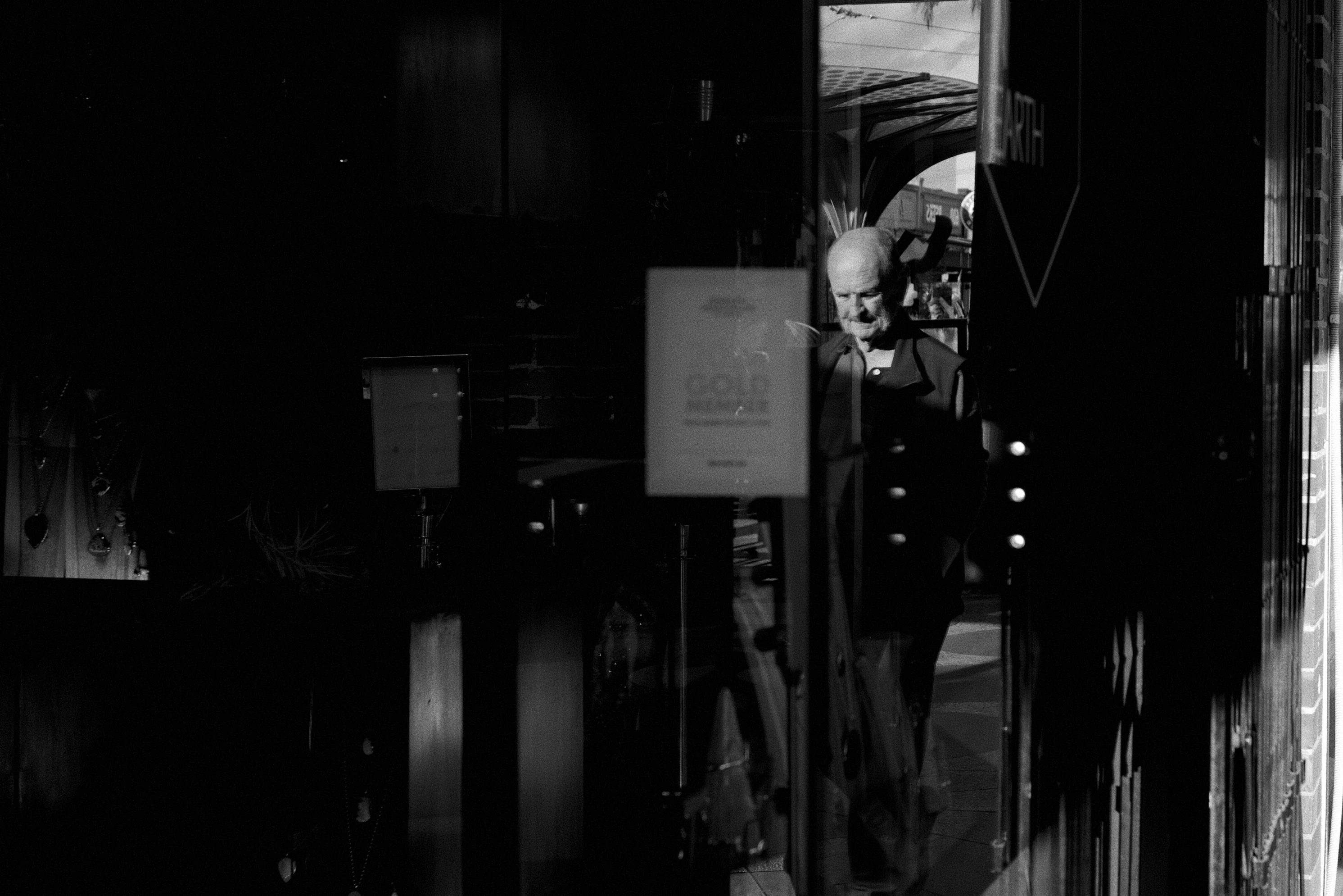 An older man, bald, is reflected on the window of a restaurant front. The photo is black and white.