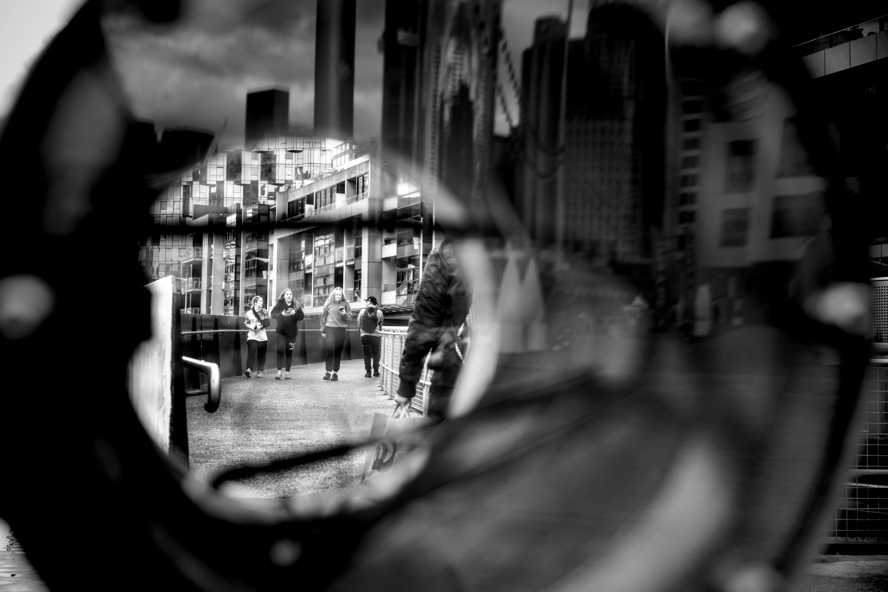 Three teenagers walks towards the camera. They are photographed through an unidentifiable round object, modern-day high-rise buildings are reflected on the edges. The photo is black and white.