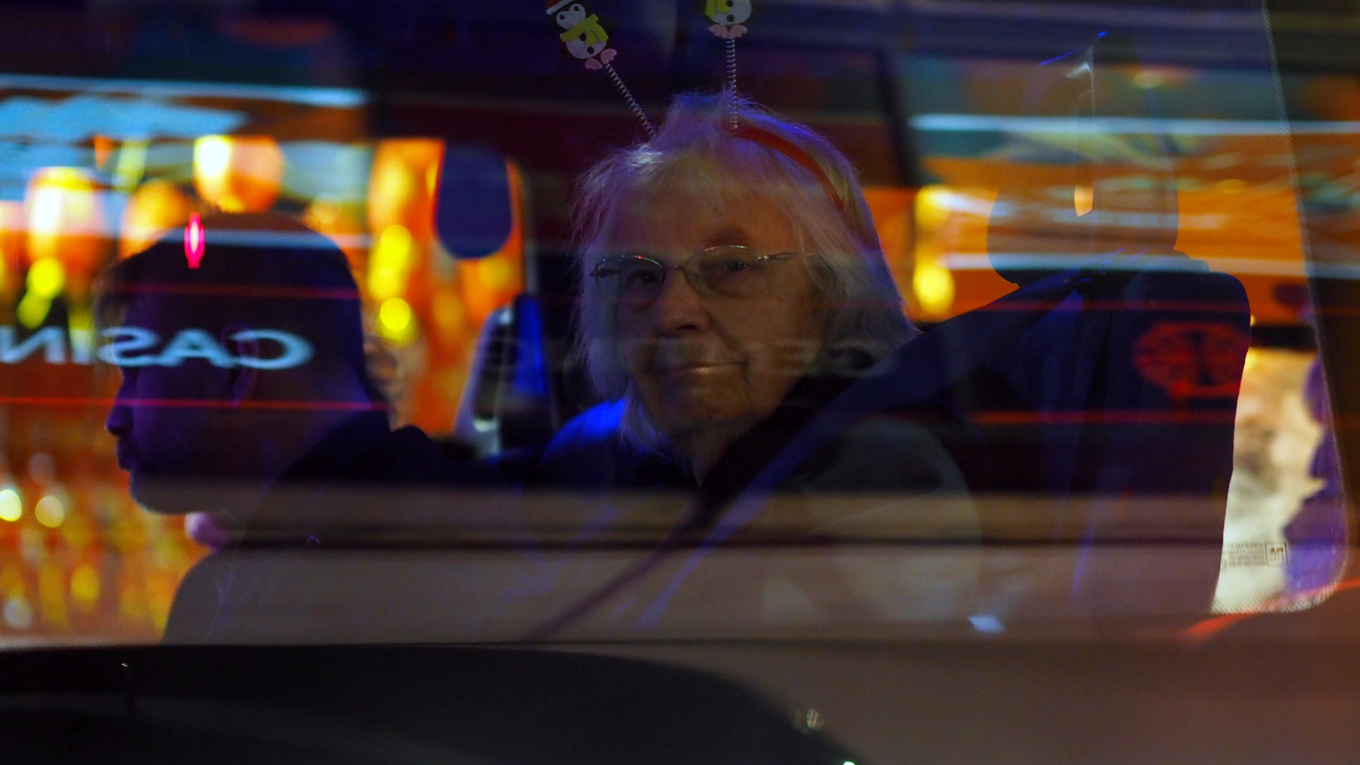 Older woman with grey hair and glasses sitting on a bus, looking straight into the camera. The windows of the bus reflect the neon signs on the street.
