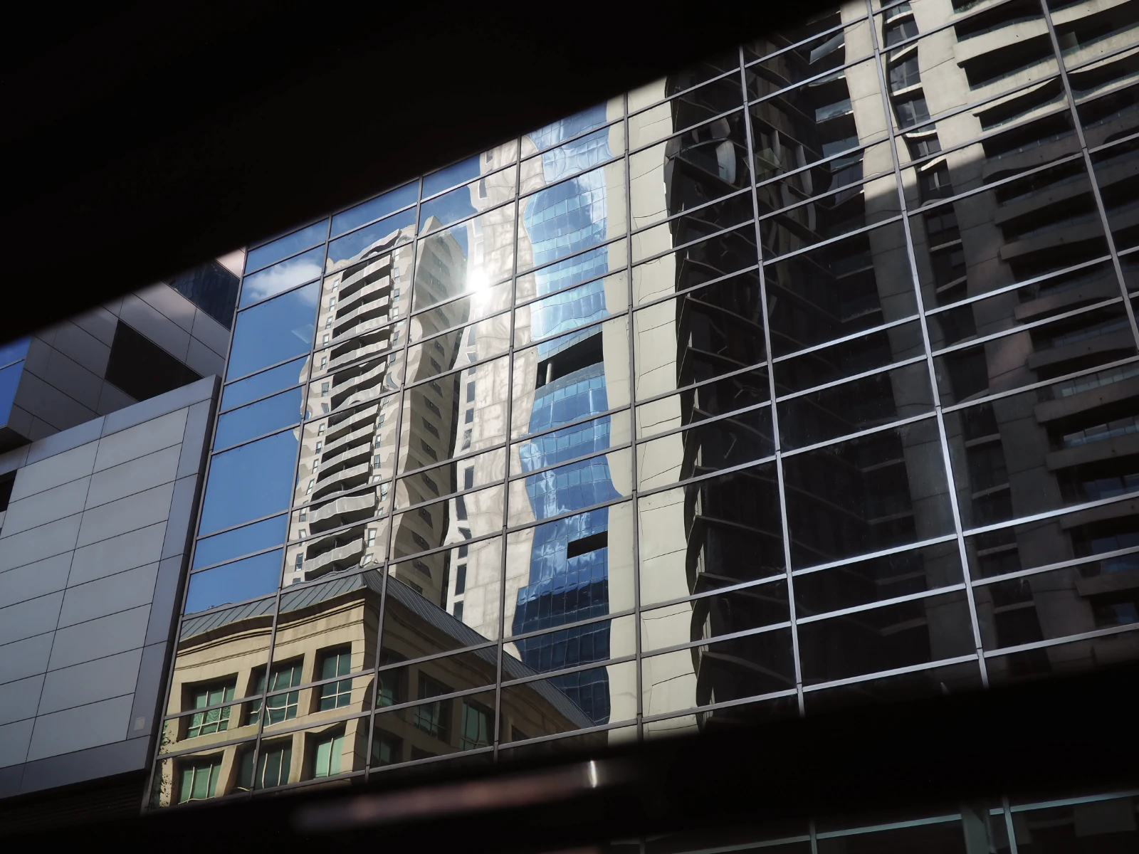 A glass-cladded building reflects the facades of buildings across the street. The view is partly blocked by the frame of the tram window.