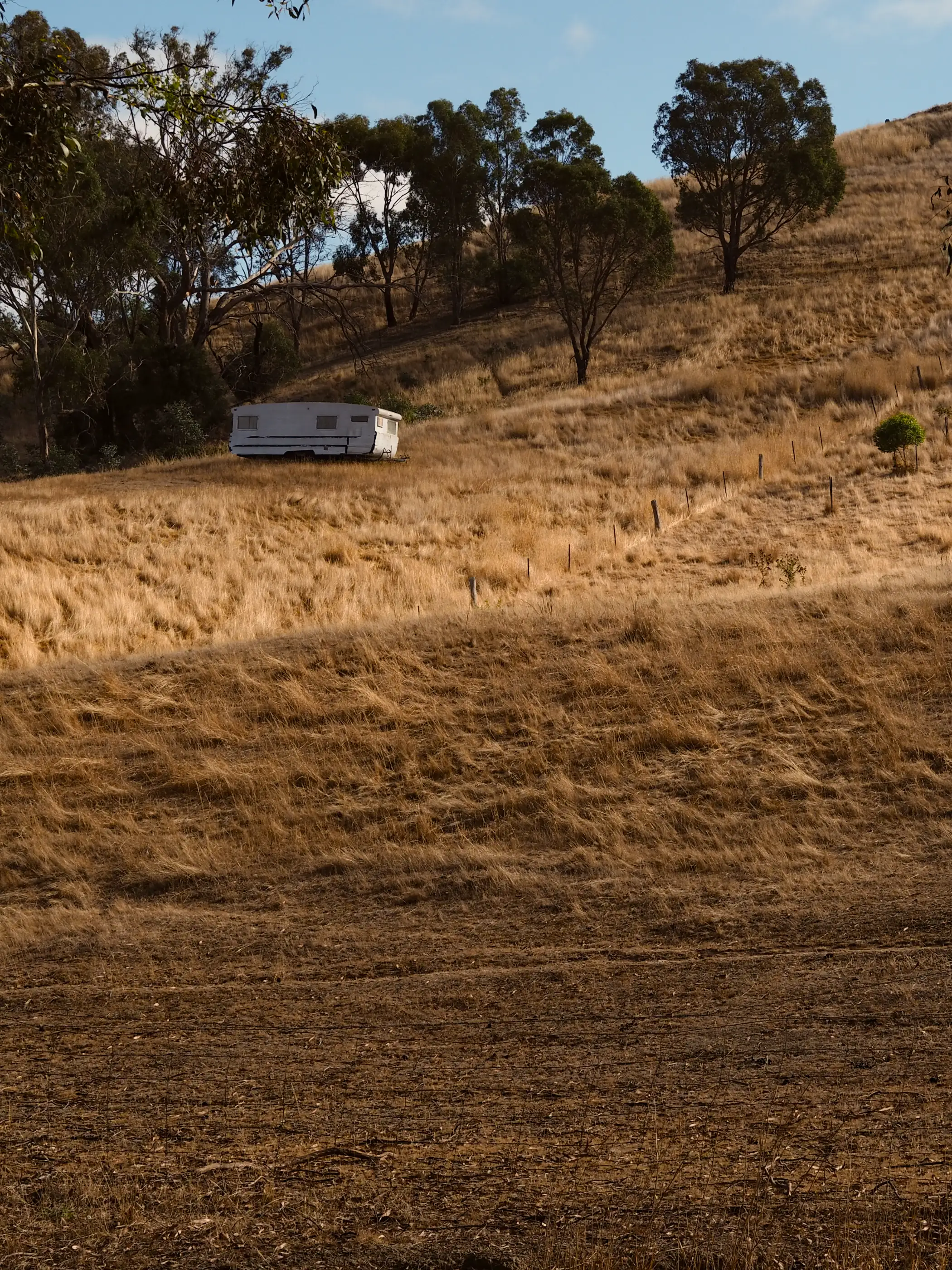 An abandoned white caravan trailer left on the slop of a hill. The hill is covered in dry, yellow gras. A couple  of trees are located in the background.