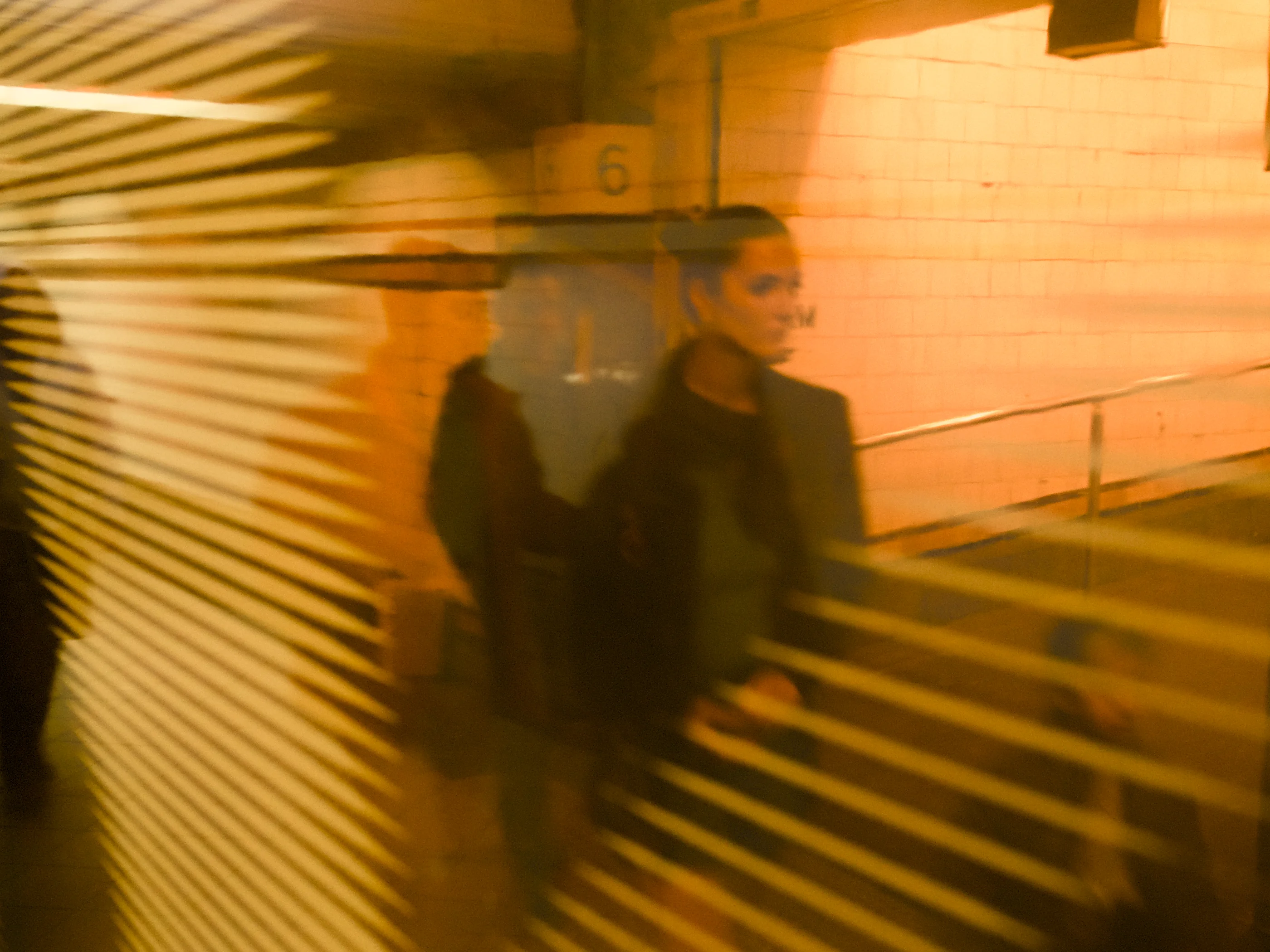 Woman and man walking through a tunnel in a train station. The pair is framed through a orange-stained plexiglass pane. In the background is a ramp leading to one of the platforms.