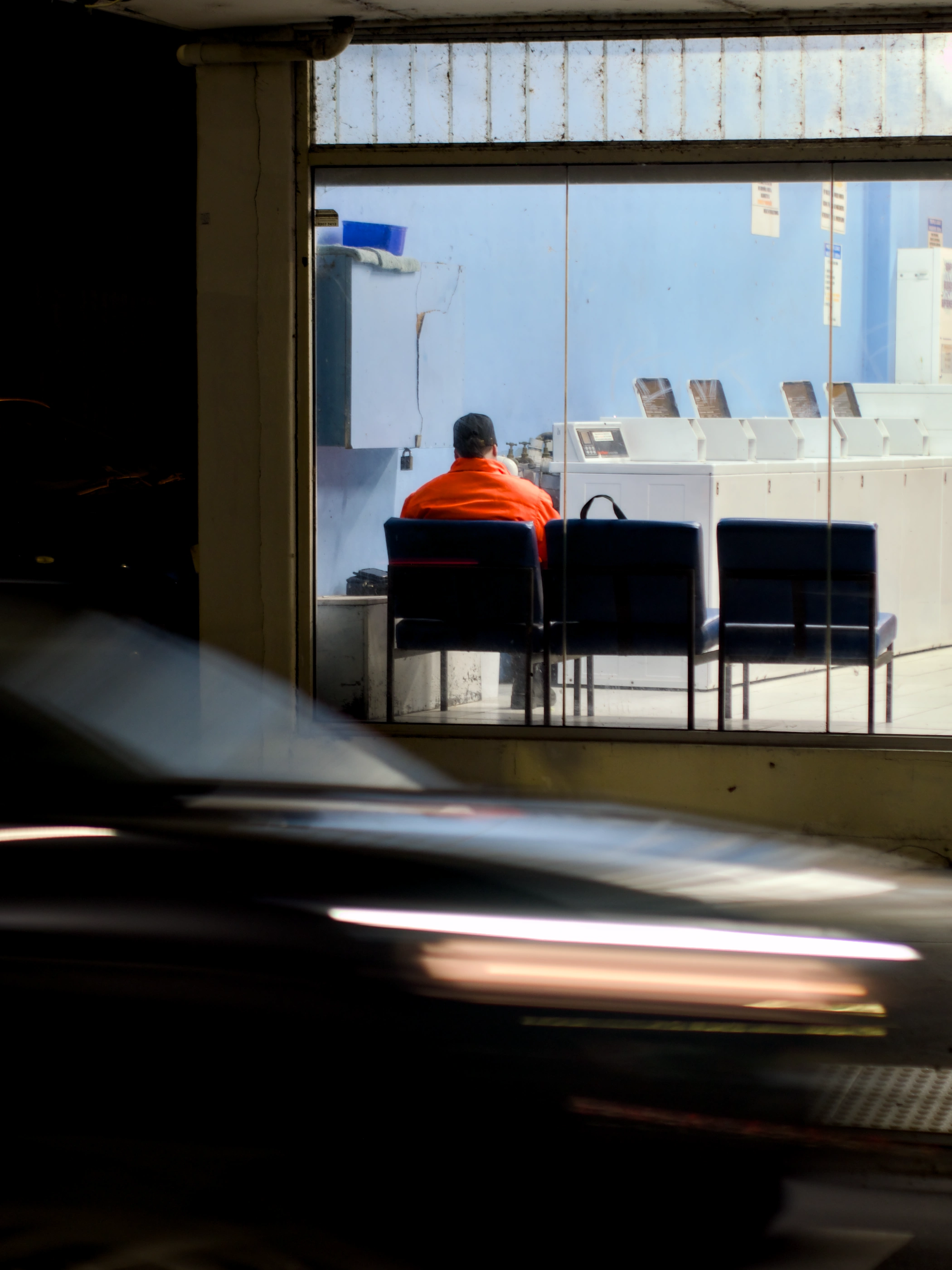 Man waring an orange jacket sitting alone inside a launderette with blue walls. In the foreground, a car passes by.