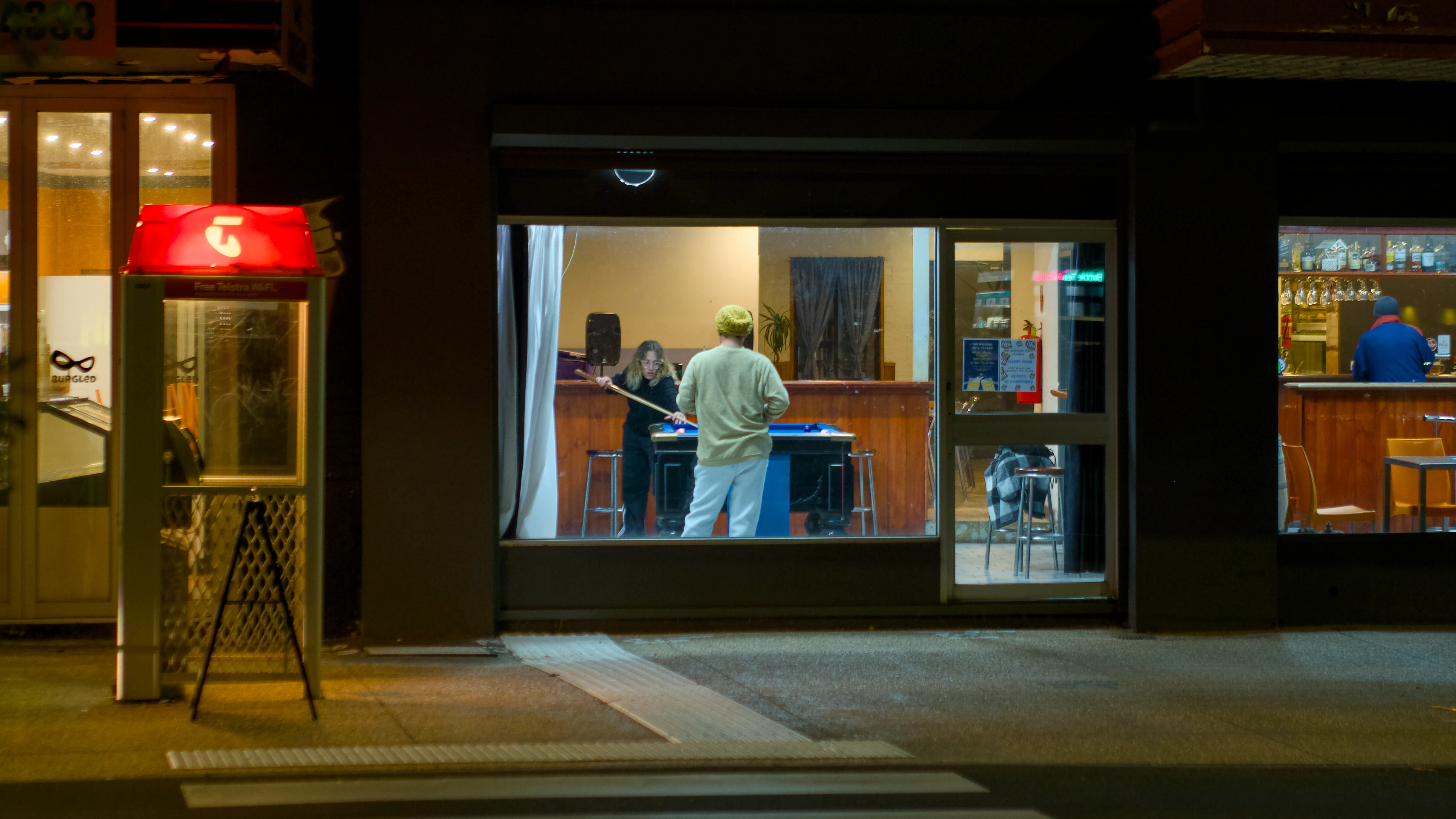 This picture combines three frames. To the left is a telephone both with a red sign in top. To the right in the window is a man in a blue jacket at the pub bar. In the centre in the window is a young couple playing pool. The scene is shot at night, with the windows illuminating the street out front.
