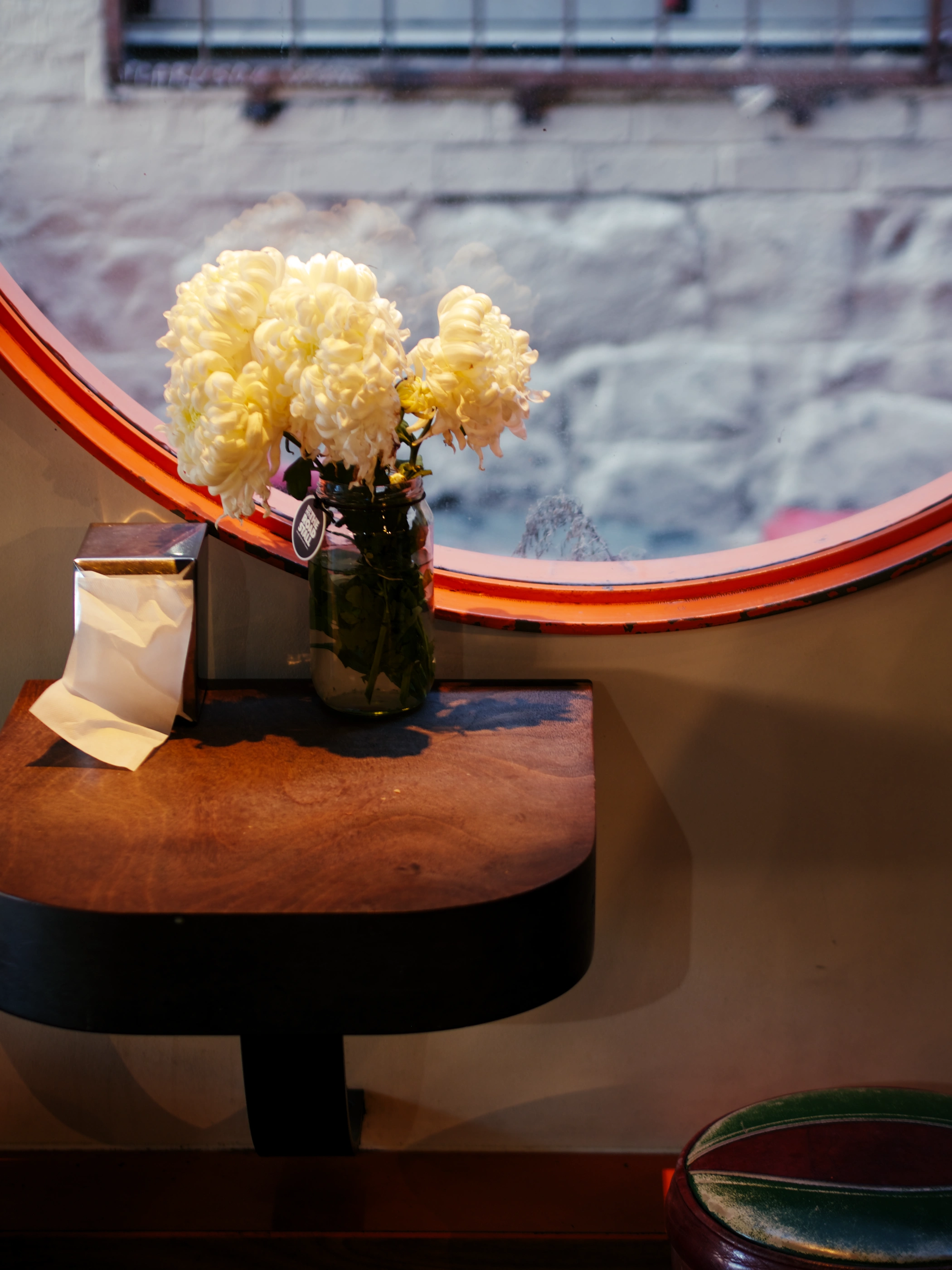 A bunch of white flowers are sitting on small wall-mounted wooden table next to a napkin dispenser. The scene is dimly lit. In the background, a circular window with an orange frame allows a look at the stone wall of the neighbouring building.
