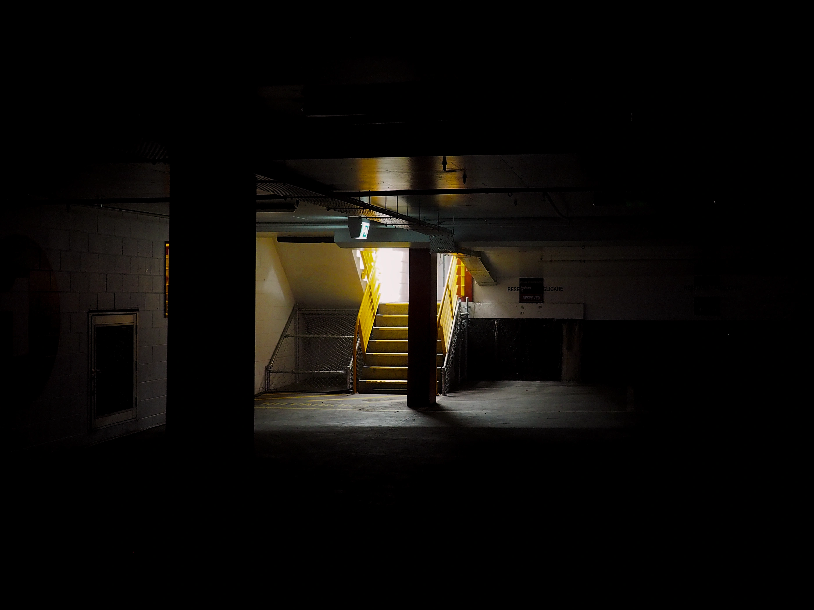 In a dark parking garage, a small set of stairs with yellow handrails leads to well-lit area.