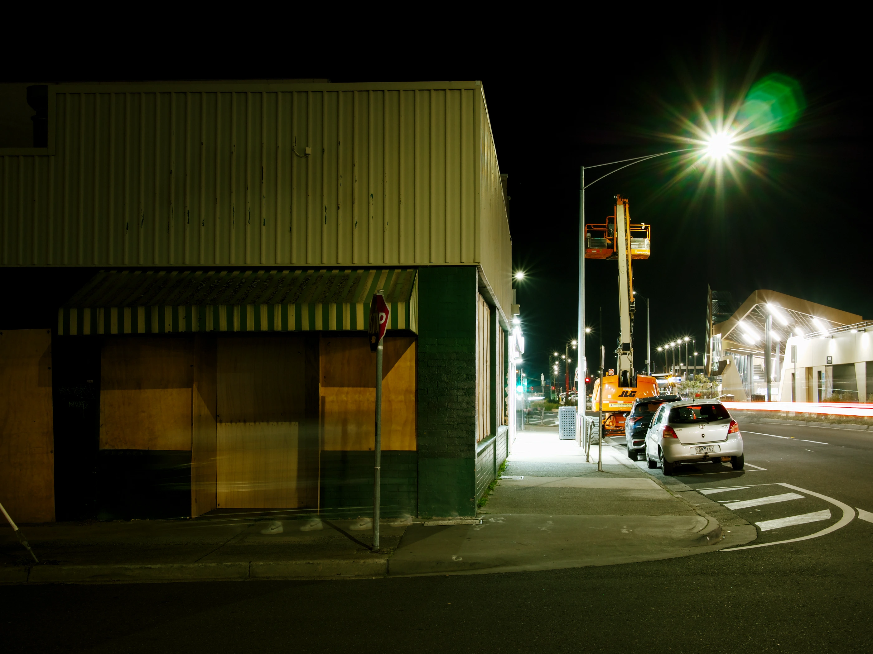 A shop front, the windows boarded up. The street around the corner is illuminated by the bright lights of a shop. In front of the boarded-up shop, you can see the shoes of a person walking past, blurred by their movement through long exposure.