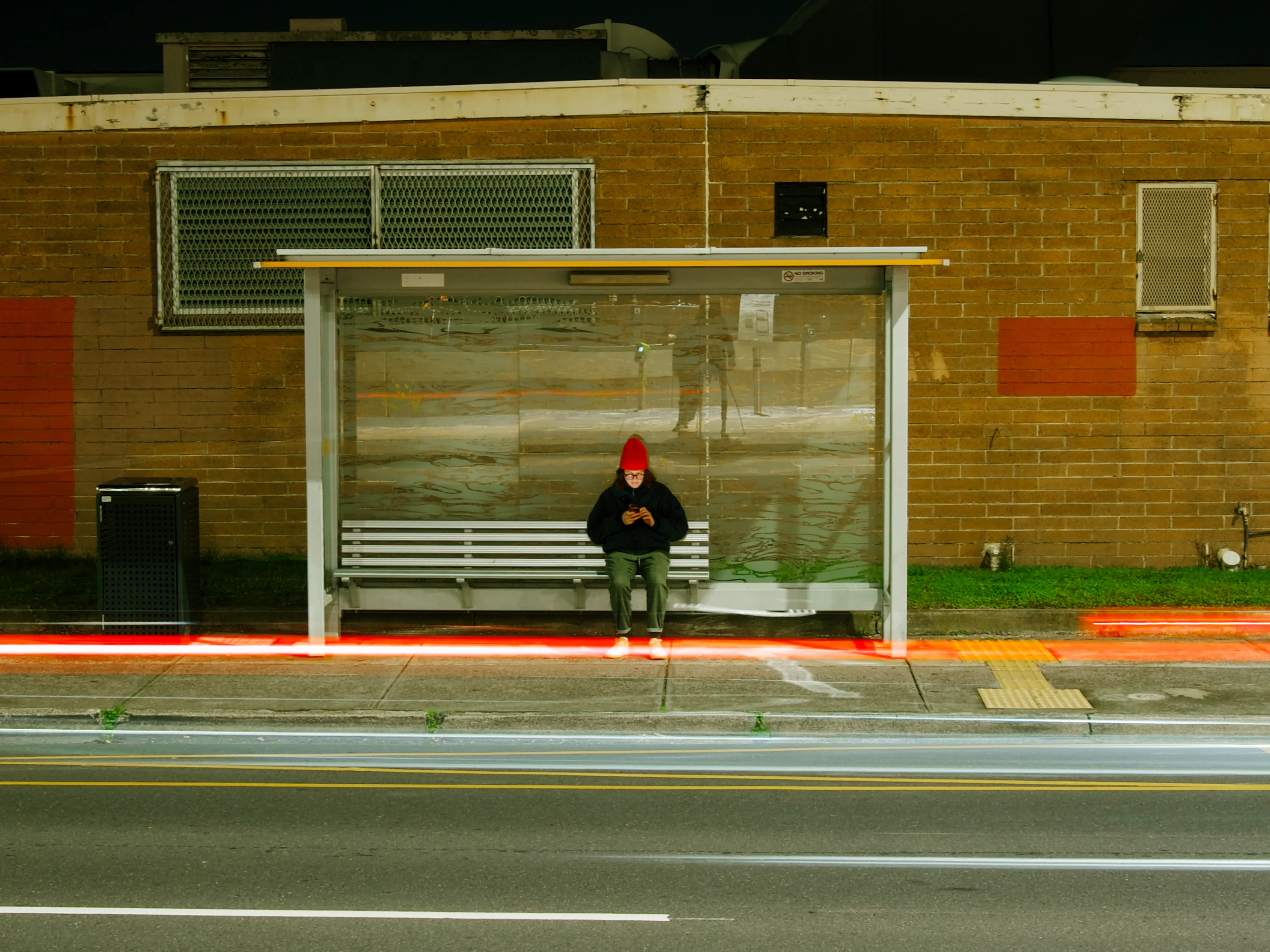 Woman wearing a red beanie sits at a bus stop at night, staring at her phone.