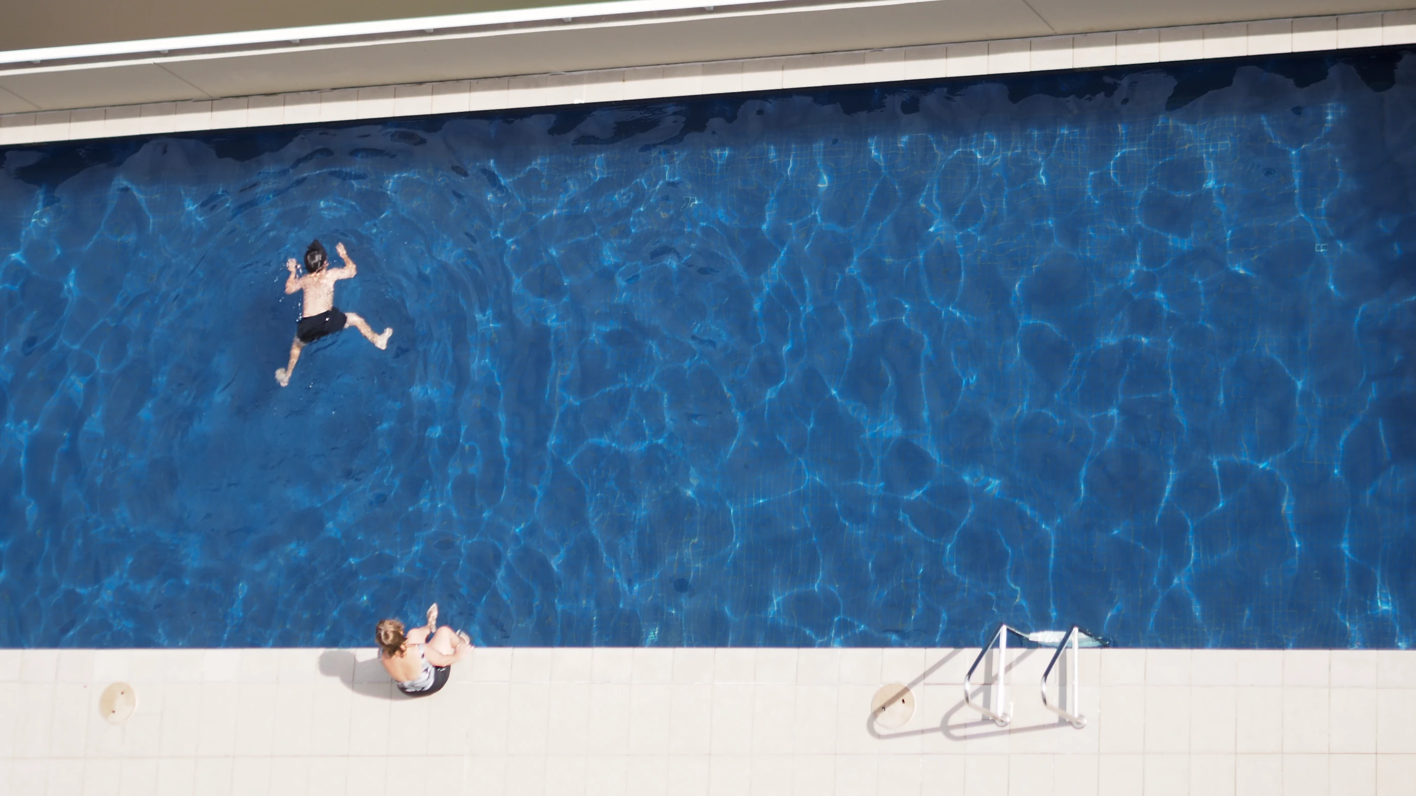 The photograph is taken from an above, showing a boy swimming in a pool with his face down. His mother sitting on the edge.