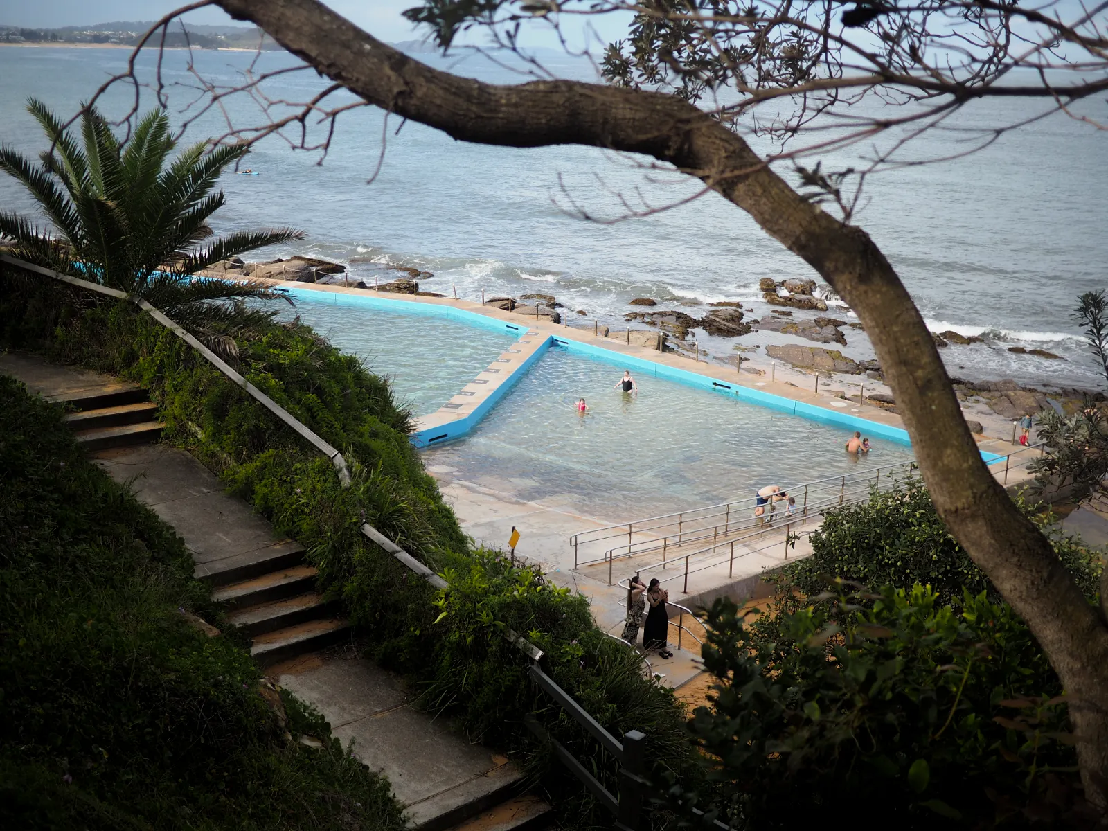 The pool is set between stairs surrounded by canopy in the foreground and the sea in background. A tree branch blocks the view in the top-left corner of the images. A few people are bathing in the pool.