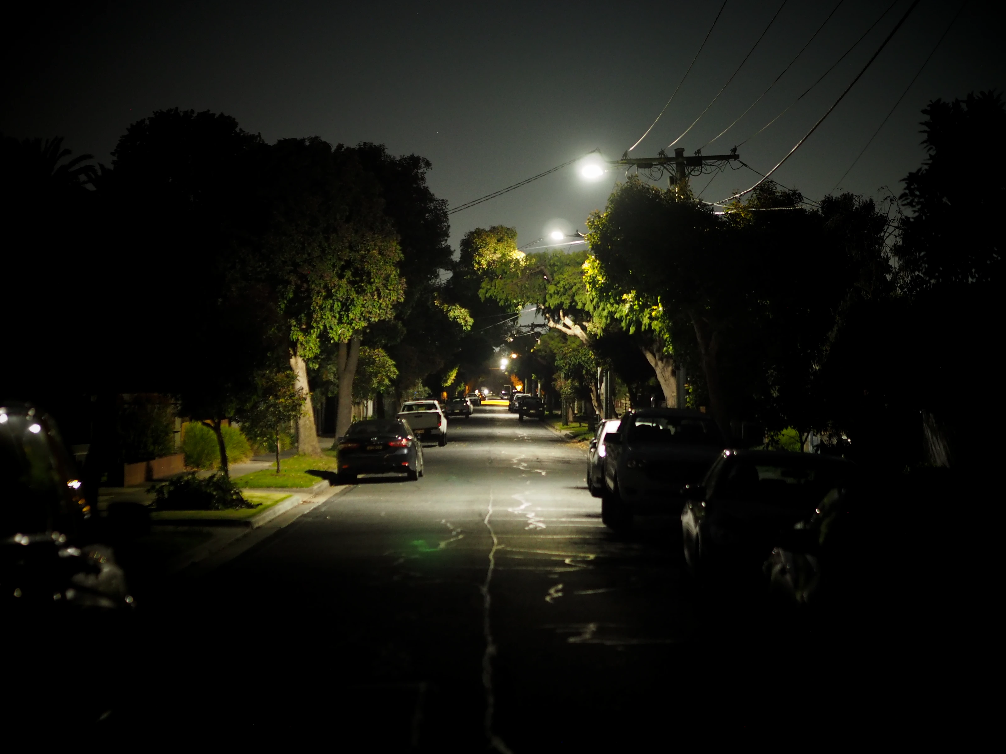 A residential road at night, photographed from a dark area to a well-lit one. Trees and cars line either side of the street.